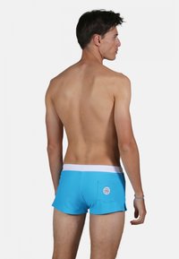 Shorts de bain bleus avec une taille blanche, fabriqués en tissu lisse. Présente une poche arrière avec un logo rond. Coupe courte, couverture minimale.