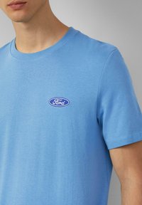 Man in een lichtblauwe T-shirt met ronde hals en een klein ovaal Ford-logo op de linkerkant van de borst, tegen een effen grijze achtergrond.