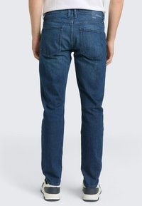 M�ężczyzna stojący, ubrany w niebieskie proste jeansy i białe sneakersy, widziany od tyłu na jednolitym tle.