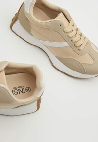 Zapatillas de tela color beige con cordones, detalles blancos y suela texturizada, mostrando vistas superior y lateral sobre un fondo blanco.