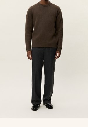 Homme portant un pull brun côtelé, un pantalon large gris foncé et des chaussures en cuir noires, debout devant un fond clair uni.