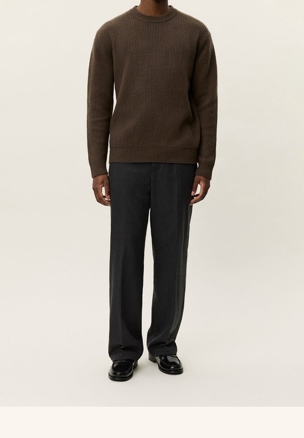 Homme portant un pull brun côtelé, un pantalon large gris foncé et des chaussures en cuir noires, debout devant un fond clair uni.