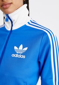 Mėlyna zip-up treniruotė striukė su balta aukšta apykakle, trimis baltomis juostelėmis ant rankovių ir balta Adidas logotipu krūtinėje. Tinklinė medžiaga.