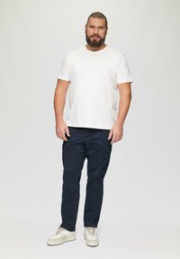 Witte katoenen T-shirt, marineblauwe cargobroek met zakken en beige sneakers. Model staat tegen een lichtgrijze achtergrond.
