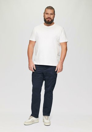 Weißes Baumwoll-T-Shirt, marineblaue Cargo-Shorts mit Taschen und beigefarbene Sneakers. Das Modell steht vor einem hellgrauen Hintergrund.