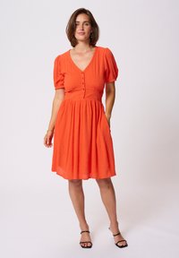 Robe orange à manches courtes avec un col en V, des boutons sur le devant et une taille froncée. Tissu doux avec une finition texturée. Sandales à lanières noires.