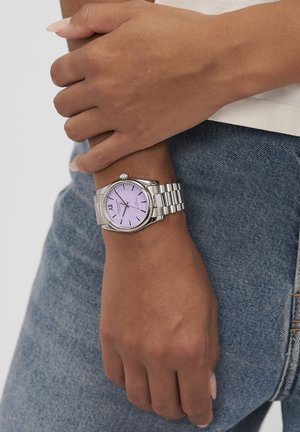 Sølvur med rund urskive og lyslilla skive. Romerske tal ved 12, 3, 6 og 9, samt et armbånd af rustfrit stål. Enkel design.