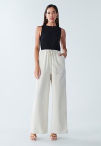 Bezroku kombinezons ar teksturētu melnu augšdaļu un gaiši bēšiem platu kāju biksēm, ar auklas jostu viduklī un sānu kabatām.