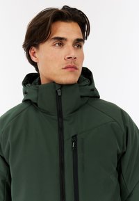 Giacca esterna verde con colletto alto, chiusura con zip e cappuccio regolabile. Presenta più tasche con zip e una trama liscia.