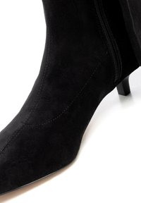 Czarny zamszowy botek za kostkę z zaostrzonym noskiem, przeszyciami, bocznym zamkiem błyskawicznym i niewielkim słupkowym obcasem. Gładka faktura i minimalistyczny design.
