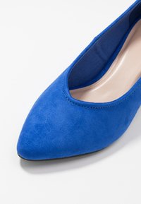 Chaussure à bout pointu en daim bleu avec une texture lisse et un détail de couture le long de l'ouverture, dotée d'une doublure intérieure douce et d'une semelle noire.