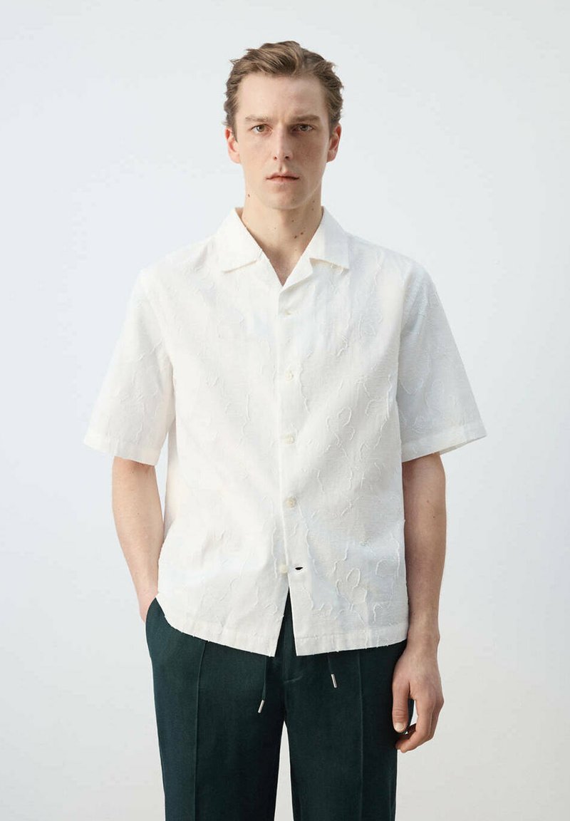 Jeune homme portant une chemise blanche texturée à manches courtes avec boutons et un pantalon foncé, debout devant un fond clair uni.