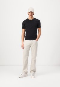 Camiseta negra de manga corta, jeans gris claro, zapatillas blancas y una gorra beige, de pie sobre un fondo blanco liso.