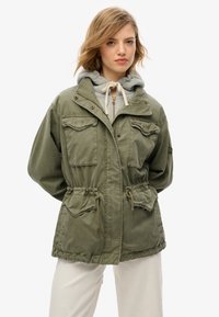 Olivgrön huva jacka med dragkedja framtill, dragsko i midjan och fyra framfickor. Bärs över en ljusgrå hoodie, vilket visar lagrade texturer.