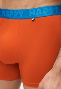 Κοντινό πλάνο ενός ατόμου που φοράει πορτοκαλί αντρικά εσώρουχα με μπλε ζώνη μέσης όπου αναγράφεται η λέξη «HAPPY» με ανάγλυφα γράμματα.