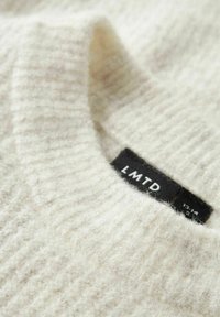 Artimu planu matyti švelnų, grietinėlės spalvos, megztos medžiagos megztinį su briaunotu tekstūru ir juodu LMNTD prekės ženklo etiketė, prisisiūta prie kaklo.