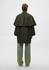 Casaco verde-oliva de grande dimensão com design de capa cropped, mangas compridas e corte descontraído, combinado com calças largas de perna larga verde-claro.