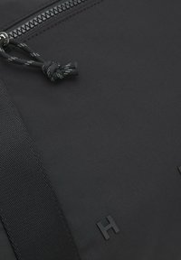 Gros plan d'un sac en tissu noir avec une sangle texturée, une fermeture éclair visible, un cordon noué et une lettre "H" embossée sur la surface.