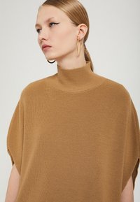 Ruskea, korkeakaulainen neule, jossa on rento istuvuus ja ribsikudottu kangas. Varustettu lyhyillä hihoilla ja sivuilla halkioilla liikkumisen helpottamiseksi.