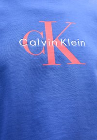 T-shirt in cotone blu con un grande logo rosso "CK" e il nome completo "Calvin Klein" in bianco, con una texture liscia e un colletto a girocollo.