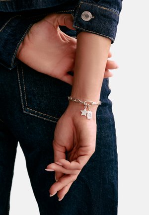 Wrist with silver chain bracelet featuring star and lock charms, hand resting behind back, wearing dark denim jeans and jacket sleeve.