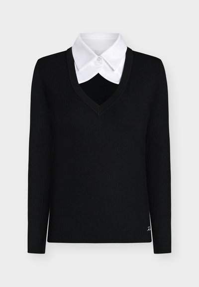 Pull noir à manches longues col en V superposé sur une chemise blanche boutonnée avec un col pointu, présenté sur un fond uni.