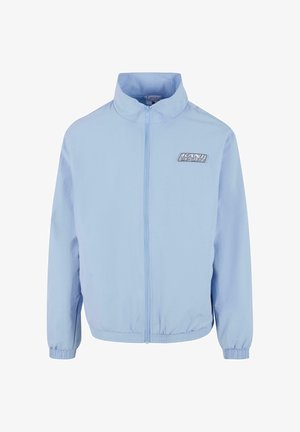 Gaiši zils apgērbs ar rāvējslēdzēju un stāvošu apkakli, elastīgām piedurknēm un apakšmalu. Uz kreisā krūškurvja ir izceltas "Kani Sport" emblēma kontrastējošā krāsā.