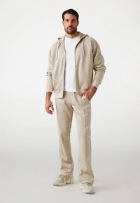 Survêtement beige comprenant un sweat à capuche zippé à motifs et un pantalon assorti avec des bandes latérales, fabriqué en tissu doux et texturé.