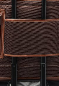 Bruine rugzak van mesh met leren accenten, voorzien van een uitschuifbare handgreep en vakken voor opbergruimte, met een gestructureerd oppervlak en stikseldetails.