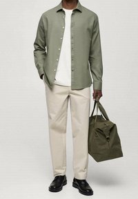 Camicia verde a maniche lunghe, t-shirt bianca, pantaloni beige, scarpe nere, mentre porta una borsa verde oliva con manici; design semplice, tessuto leggero.