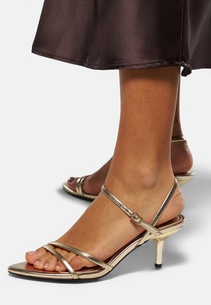 Guldiga sandaletter med låg stiletto-häl, med en metallisk finish, öppen tå-design och justerbar ankelrem. Slät textur.