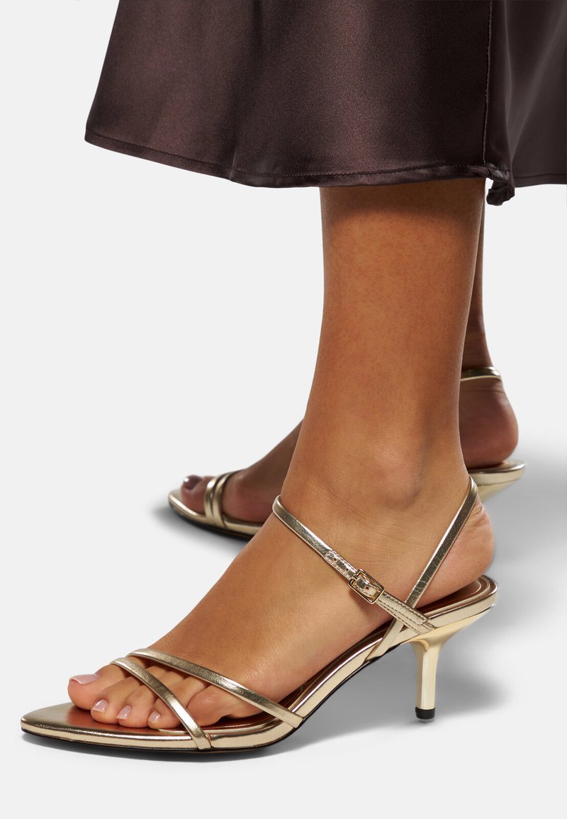 Guldiga sandaletter med låg stiletto-häl, med en metallisk finish, öppen tå-design och justerbar ankelrem. Slät textur.
