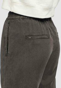 Close-up van donkergrijze elastische taillebroek met achterzak en klein merklabel, gedragen met een witte top.