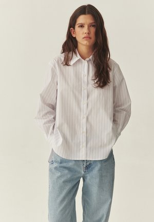 Young person with long dark hair wearing a white pinstriped button-up shirt and light blue jeans, hands in pockets, neutral background.