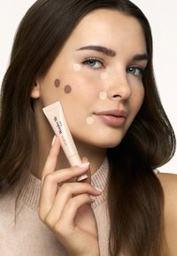 Hellbeige 3-in-1-Cremetube, gehalten von einer Person mit einer glatten Haut, die Punkte verschiedener Foundation-Töne auf ihrer Wange zeigt.