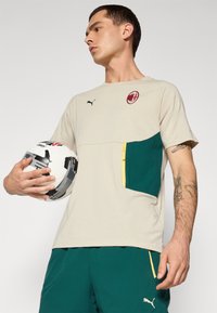Beige t-shirt med grønne detaljer i lommen og gul lynlås, der har et Puma-logo og et rødt klubemblem, iført af en person, der holder en fodbold.