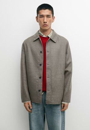 Jeune homme portant une veste taupe boutonnée par-dessus un pull rouge et une chemise beige avec un jean bleu, debout devant un fond blanc uni.