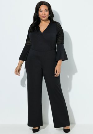 Kurvige Frau steht selbstbewusst da, trägt einen schwarzen Overall mit Spitzendetails an dreiviertellangen, ausgestellten Ärmeln und schwarze Absätze.