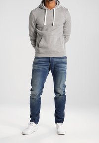 Grå huvtröja med vita snoddar, framficka, tillsammans med blå urblekta jeans och vita sneakers, stående mot en ljus bakgrund.