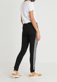 Svarta sweatpants med vita ränder längs sidorna, åtsittande vid anklarna, kombinerade med en enkel vit t-shirt och bruna sneakers.