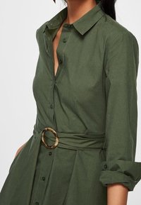 Femme portant une robe boutonnée vert olive avec des manches retroussées et une ceinture assortie avec une boucle en écaille de tortue circulaire.