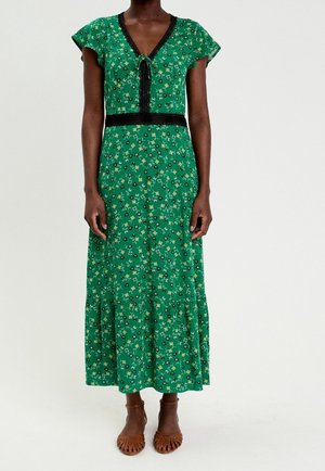 Kvinde iført en grøn blomster-midi-kjole med sort blondekant, flutterærmer og brune flettede sandaler, stående mod en hvid baggrund.