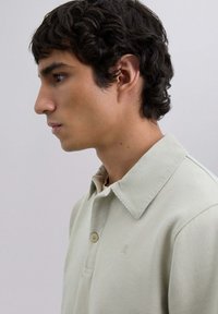 Camisa polo verde claro con un tejido texturizado, un cuello de canalé, dos botones y un pequeño logo en el pecho. Perfil lateral de un modelo.