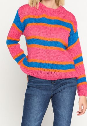 Frau trägt einen pinken gestrickten Pullover mit blauen und orangefarbenen horizontalen Streifen, kombiniert mit blauen Jeans, steht mit einer Hand in der Hüfte.