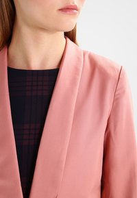 Ljusrosa blazer i slät tyg med slagkrage. Under jackan finns en marinblå klänning med ett rutigt mönster i mörkröd och marinblå.