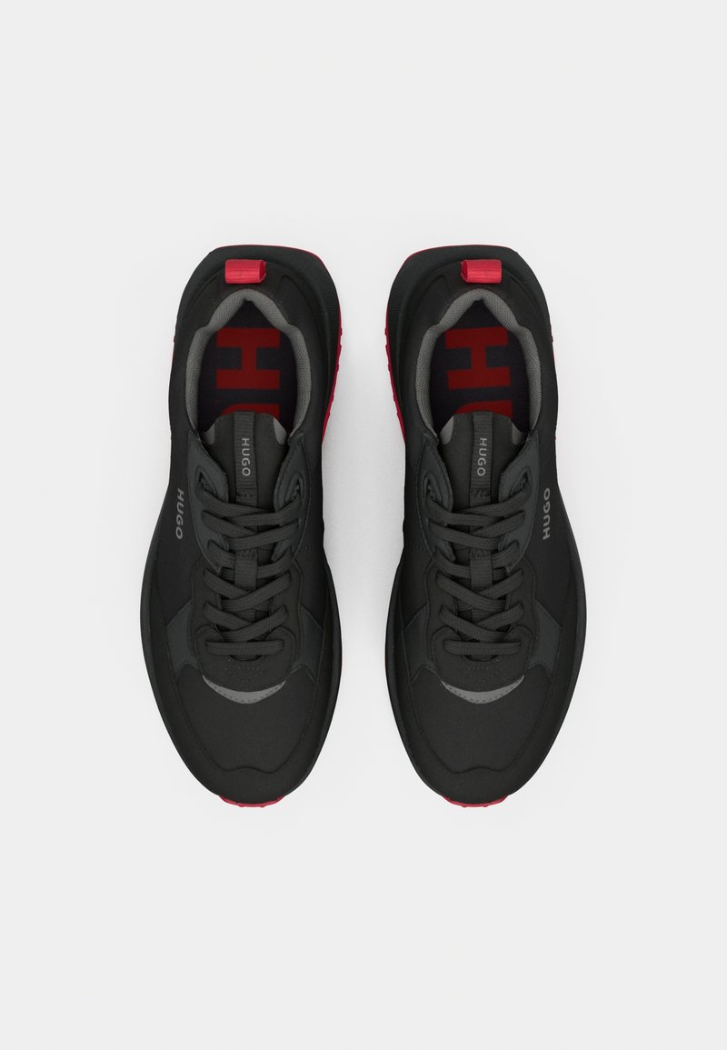 Schwarze Sportschuhe mit einem strukturierten Obermaterial, grauen Akzenten und roten Details auf der Sohle. Sie verfügen über flache Schnürsenkel und eine gepolsterte Innensohle.