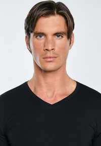 Noor mees keskmise pikkusega pruunide juustega ja siniste silmadega, kes kannab musta V-kaelusega särki, seisab vastamisi ühevärvilise heleda tausta ees.