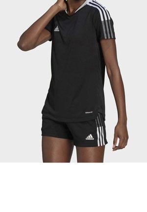 Osoba v černém sportovním oblečení Adidas s bílými pruhy na rukávech a šortkách, stojící před jednobarevným pozadím.