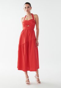 Punane kaela taha seotud kleit, millel on istuv bodi, keerutatud disain ja lõhikuga detail. Õhuline, keskmise pikkusega seelik kihilise tekstuuriga. Kandes valgeid kontsakingi.