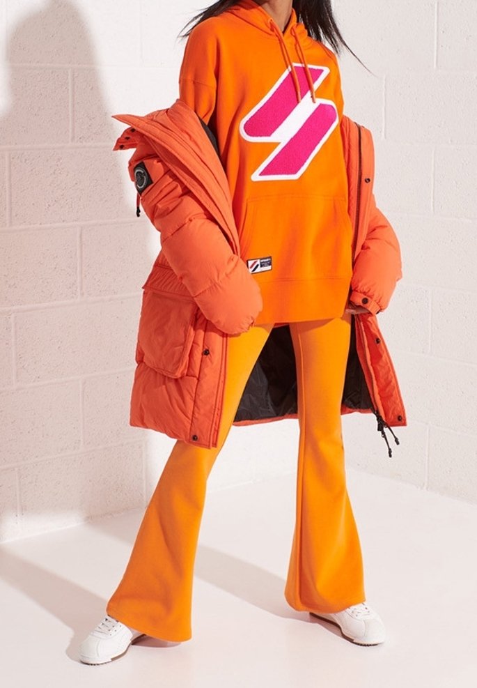 Chaqueta acolchada naranja sobre una sudadera con capucha naranja con un logo rosa, combinada con pantalones de campana naranjas y zapatillas blancas. Textura suave y colores vibrantes.